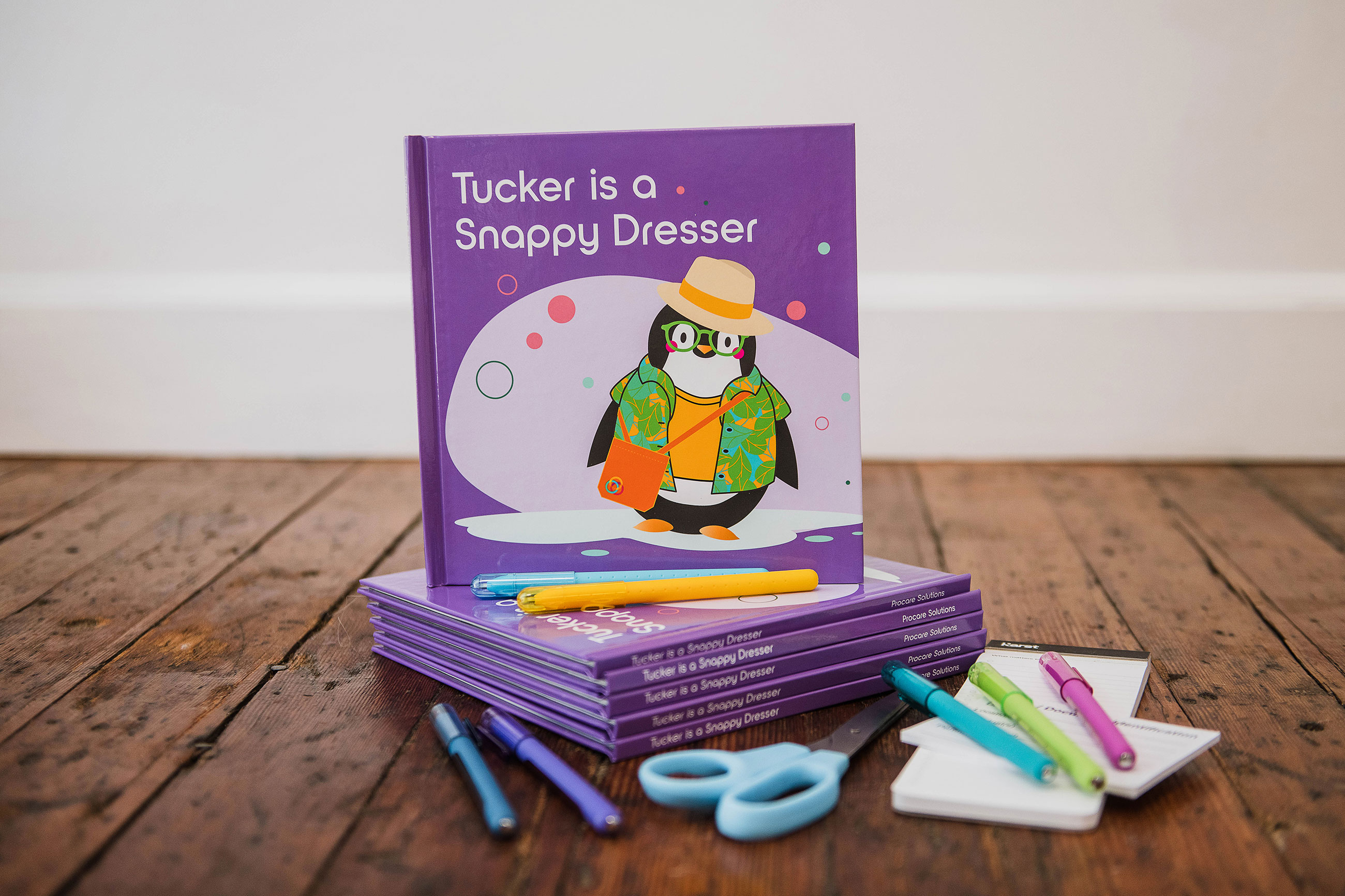 Photo of the printed children's books, Tucker is a Snappy Dresser. A hard cover book is propped up on a stack of books with classroom supplies in the foreground.