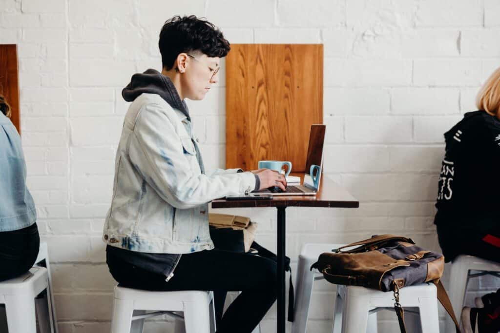 A person with short hair and glasses wearing a denim jacket works on a laptop in a coffee shop.